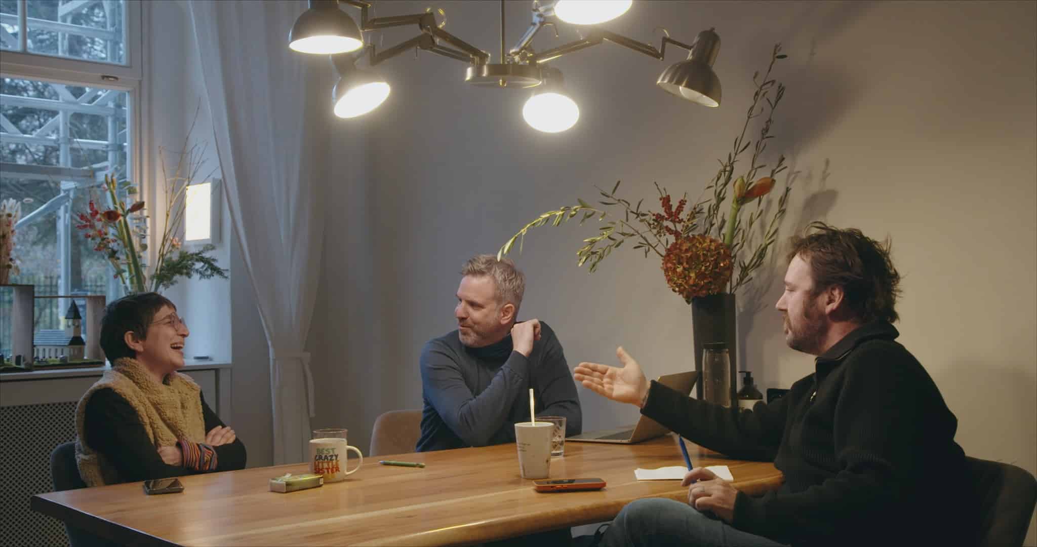 Zwei Männer mittleren Alters sitzen und eine ältere Frau sitzen an einem Tisch und unterhalten sich. Sie lacht.